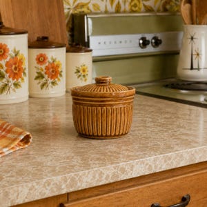 May include: A small, round, brown ceramic jar with a matching lid. The jar features a vertical ribbed texture along its sides and a circular, ridged pattern on the top of the lid. The glaze has a glossy, warm brown finish with darker tones.