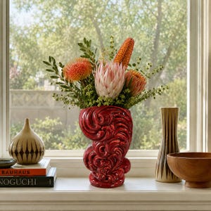 May include: A glossy red ceramic vase with a swirling, organic relief pattern holds a bouquet of orange and pink protea flowers and green foliage. The vase sits on a windowsill next to a tan patterned vase, a wooden bowl, and two books.
