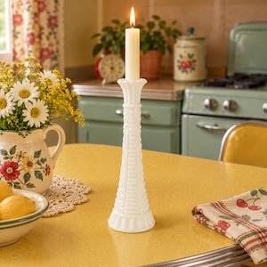 Può includere: Un alto candelabro in vetro opalino bianco con un motivo geometrico strutturato e una base svasata. Una singola candela affusolata color crema è accesa e inserita nella parte superiore, diffondendo una luce calda con la sua piccola fiamma.