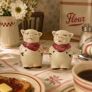 May include: Two ceramic pig figurines standing side by side, each featuring a glossy cream-colored finish, a painted dark red scarf around the neck, and small brown accents on the feet. The pigs have closed eyes and a serene expression.