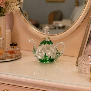 May include: A clear glass ring holder shaped like a miniature teapot, featuring green glass leaves and white floral accents at the base. A gold-toned ring with a clear stone is placed on the vertical stem of the teapot, which serves as a post.