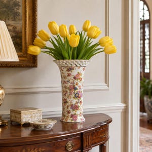 May include: A tall, cylindrical porcelain vase with a flared, lattice-patterned rim, decorated with a colorful floral pattern of pink, blue, and yellow blossoms on a white background, filled with a large bouquet of bright yellow tulips.