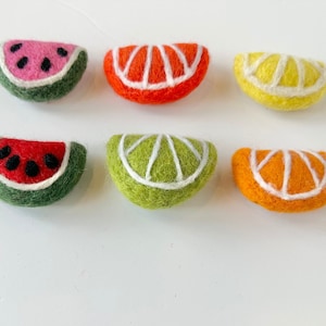 May include: Six felt fruit slices, including watermelon, orange, and lime, arranged on a white surface with a wooden ruler for scale.