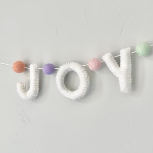 May include: A white felt garland spelling the word JOY in capital letters, interspersed with small felt balls in pastel shades of peach, purple, pink, and mint green, all strung together on a thin white cord against a plain light background.