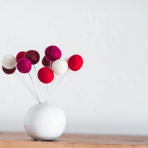 May include: A white ceramic vase with a round base holds a bouquet of felt balls in shades of red, pink, and white.