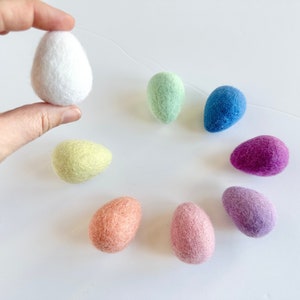 May include: A set of eight felt Easter eggs in various pastel colors, including white, light green, blue, purple, pink, and yellow. The eggs are arranged in a circle on a white surface.