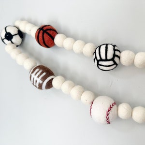 May include: A white felt garland with felt balls and different sports balls including a soccer ball, basketball, volleyball, football, and baseball.