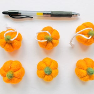 May include: Six felt orange pumpkins with green stems and white string loops. The pumpkins are arranged in two rows of three on a white surface.