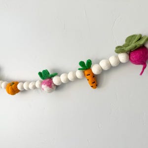 May include: A felt garland featuring various vegetables, including carrots, radishes, and a pumpkin, interspersed with white felt balls. The vegetables are in shades of orange, pink, and green, creating a colorful and whimsical decorative piece.