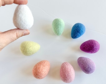 May include: A set of eight felt Easter eggs in various pastel colors, including white, light green, blue, purple, pink, and yellow. The eggs are arranged in a circle on a white surface.