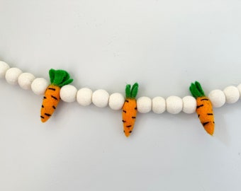 May include: A decorative garland featuring a string of round, white felted balls interspersed with three orange felt carrots. Each carrot has a green felt top and black stitched horizontal lines, creating a repeating pattern along the string.