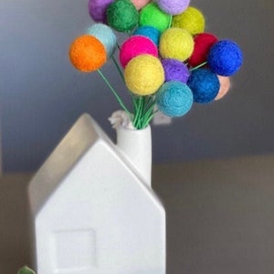 May include: A white ceramic house-shaped vase with a chimney holds a bouquet of colorful felt balls. The balls are in shades of blue, green, yellow, orange, pink, and purple.
