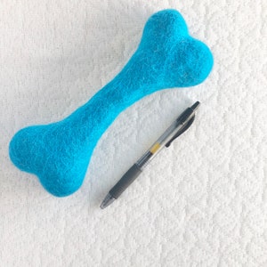 May include: A bright blue felt dog bone toy with a pen lying next to it on a white surface.