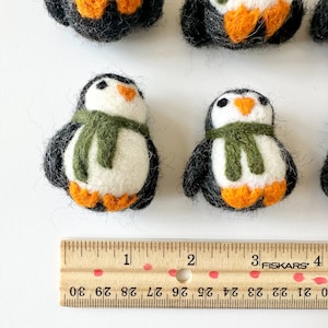 May include: Five felt penguin ornaments, two with green scarves, on a white surface with a wooden ruler marked in inches. The penguins are approximately 2 inches tall.