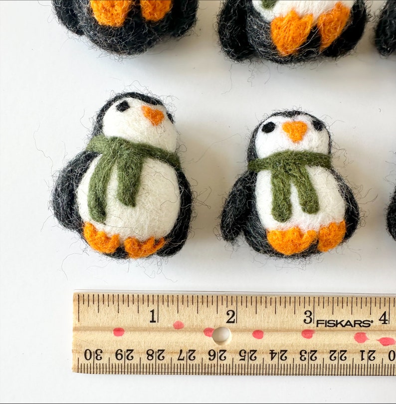 May include: Five felt penguin ornaments, two with green scarves, on a white surface with a wooden ruler marked in inches. The penguins are approximately 2 inches tall.