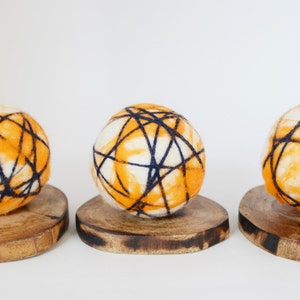May include: Three orange and white felted balls with black yarn woven through them, displayed on wooden bases.