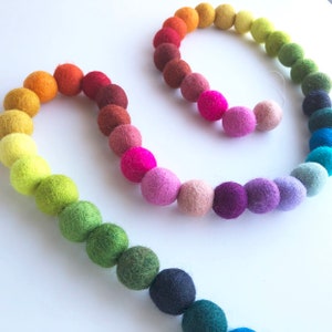 May include: A rainbow garland made of felt balls. The garland is strung together with a thin white thread. The felt balls are in a variety of colors, including red, orange, yellow, green, blue, purple, and pink.