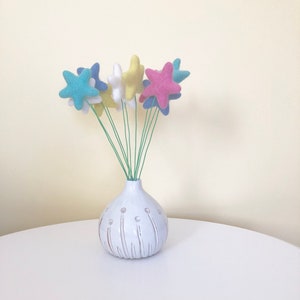 May include: A white ceramic vase with a textured surface and a bunch of felt star-shaped flowers in blue, yellow, and pink.