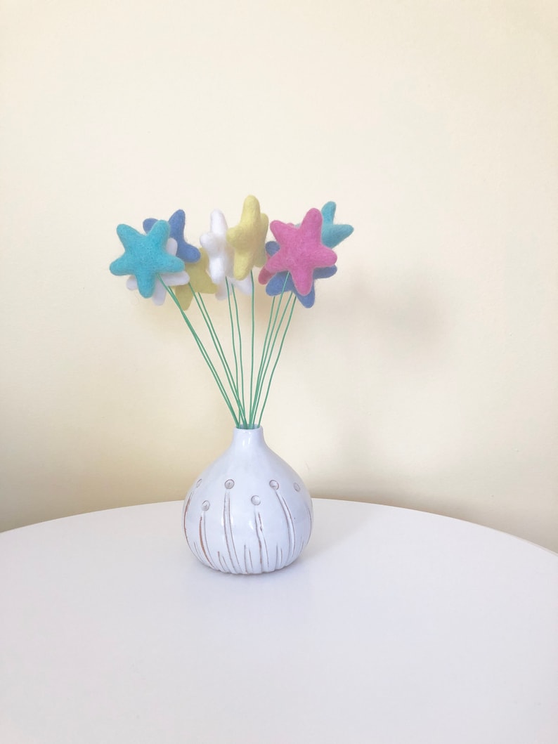 May include: A white ceramic vase with a textured surface and a bunch of felt star-shaped flowers in blue, yellow, and pink.