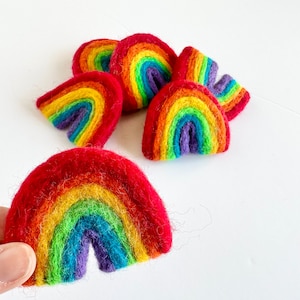 May include: A set of felt rainbow ornaments in various shades of red, orange, yellow, green, blue, indigo, and violet. The rainbows are arranged in a pile, with one rainbow held in a hand.
