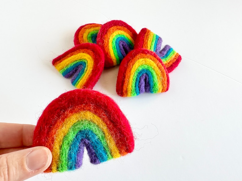 May include: A set of felt rainbow ornaments in various shades of red, orange, yellow, green, blue, indigo, and violet. The rainbows are arranged in a pile, with one rainbow held in a hand.