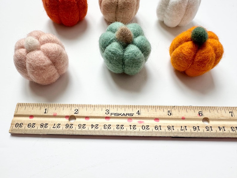 May include: Six felt pumpkins in various colors, including orange, green, white, and pink, are arranged on a white surface. A wooden ruler with inch markings is placed below the pumpkins.