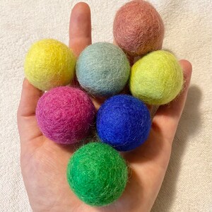 May include: A hand holding seven colorful felt balls. The balls are in shades of pink, red, yellow, green, blue, and light blue.