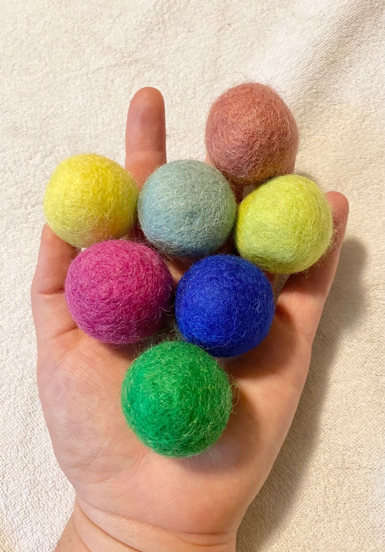May include: A hand holding seven colorful felt balls. The balls are in shades of pink, red, yellow, green, blue, and light blue.