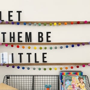 May include: A white wall with three shelves displaying the phrase "Let them be little" in black letters on clear acrylic blocks. The shelves are decorated with colorful pom-pom garlands.