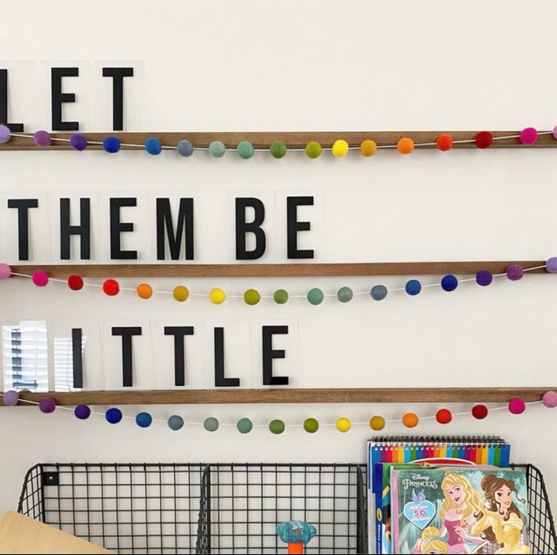 May include: A white wall with three shelves displaying the phrase "Let them be little" in black letters on clear acrylic blocks. The shelves are decorated with colorful pom-pom garlands.