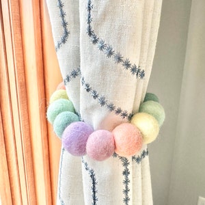 May include: A pastel rainbow colored felt ball curtain tieback. The tieback is made of a string of felt balls in various shades of pink, green, and yellow. The tieback is tied around a white curtain with a black embroidered pattern.