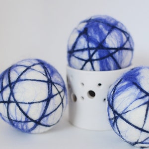 May include: Three white and blue decorative spheres with a dark blue web-like design. One sphere rests on a white ceramic holder with circular cutouts. The spheres have a soft, textured appearance, suggesting a felted material.