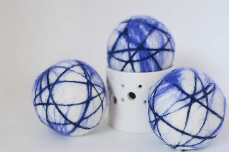 May include: Three white and blue decorative spheres with a dark blue web-like design. One sphere rests on a white ceramic holder with circular cutouts. The spheres have a soft, textured appearance, suggesting a felted material.