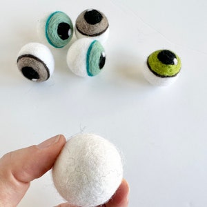 May include: A collection of felt eyeballs with different colored irises and pupils. The eyeballs are white with brown, green, and black details. One white felt eyeball is being held by a hand.