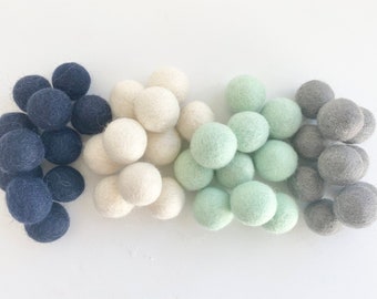 May include: A collection of felt balls in four colors: dark blue, white, light green, and gray. The balls are arranged in four rows, with the dark blue balls on the left, the white balls in the middle, the light green balls on the right, and the gray balls on the far right.