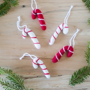May include: Five felt candy cane ornaments with red and white stripes hanging from white twine on a wood background. The ornaments are arranged in a semi-circle with the tips of the candy canes pointing towards the center.