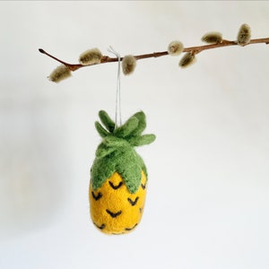 May include: A felt pineapple ornament with a green top and yellow body with black detailing. The ornament is hanging from a branch with fuzzy buds.