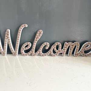 May include: Wooden "Welcome" sign in cursive script. The letters are white with a border of small, light brown spheres. The sign is placed on a white surface against a gray wall, creating a welcoming home decor accent.