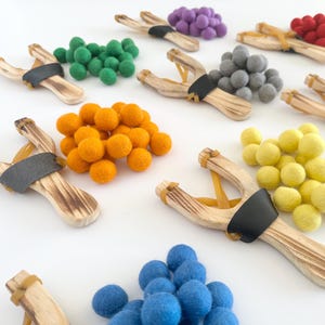 May include: Wooden slingshots with black leather rests are paired with colorful felt balls in green, orange, yellow, blue, gray, purple, and red. The slingshots have a natural wood grain finish and yellow rubber bands.