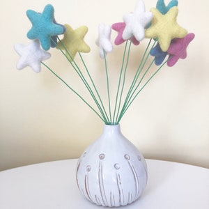 May include: A white ceramic vase with a textured surface and a round shape holds a bouquet of felt star-shaped flowers in various colors, including blue, yellow, pink, and white.
