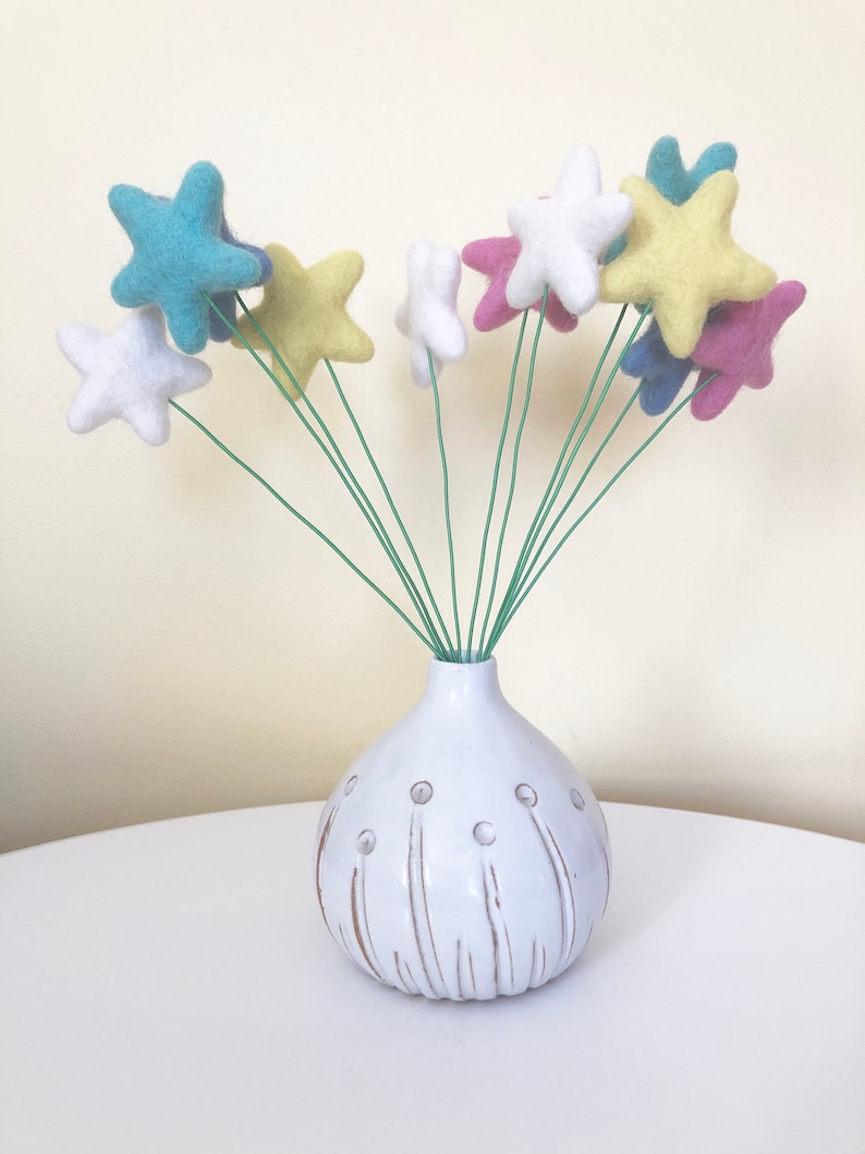 May include: A white ceramic vase with a textured surface and a round shape holds a bouquet of felt star-shaped flowers in various colors, including blue, yellow, pink, and white.