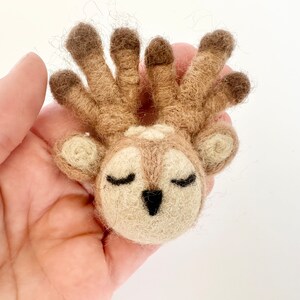 May include: A small, hand-felted deer with closed eyes and a brown and white color scheme. The deer has a large, fluffy head with a set of antlers.