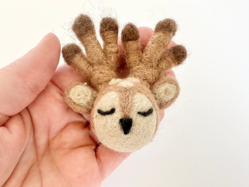 May include: A small, hand-felted deer with closed eyes and a brown and white color scheme. The deer has a large, fluffy head with a set of antlers.