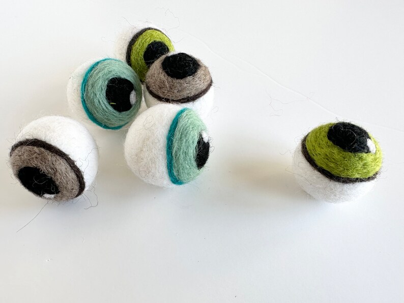 May include: Five white felt balls with different colored eyes. The eyes are green, brown, and blue. The balls are all about the same size and are arranged in a cluster.