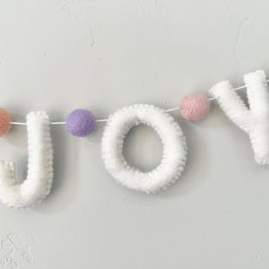 May include: A decorative garland featuring the white felt letters J, O, and Y strung on a thin white cord. The letters are interspersed with small, round felt balls in pastel shades of peach, lavender, dusty pink, and mint green.