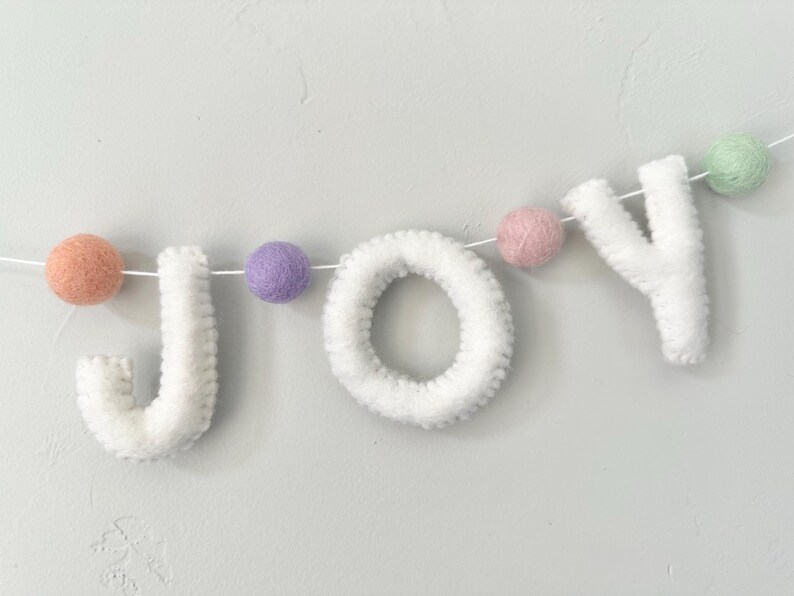 May include: A decorative garland featuring the white felt letters J, O, and Y strung on a thin white cord. The letters are interspersed with small, round felt balls in pastel shades of peach, lavender, dusty pink, and mint green.