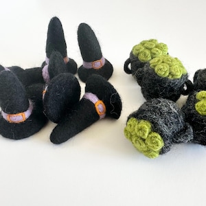 May include: A set of felt witch hats and felt cauldron ornaments. The hats are black with purple and orange accents. The cauldrons are black with green felt rosettes.