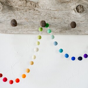 May include: A rainbow felt ball garland with a gradient of colors from red to purple. The garland is strung on white thread and is hanging from a piece of driftwood.