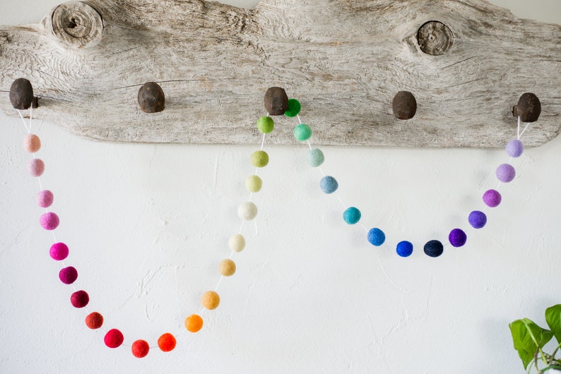 May include: A rainbow felt ball garland with a gradient of colors from red to purple. The garland is strung on white thread and is hanging from a piece of driftwood.