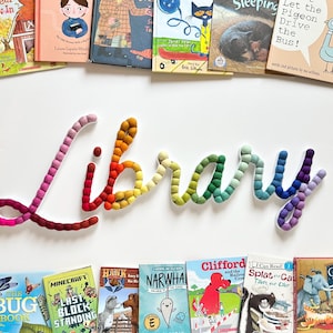 May include: A rainbow-colored felt word "Library" with white backing. The word is made of small, round felt balls. The word is surrounded by a variety of children's books.